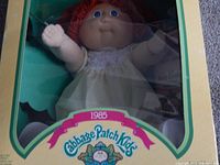 Front view of boxed 1985 Cabbage Patch Kids doll showing red hair, blue eyes, white dress, and packaging with brand and year.