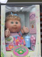 Front view of boxed Cabbage Patch Kids baby doll with visible accessories and Birth Certificate and Adoption Papers inside the box