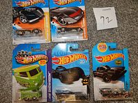 Photo of five Hot Wheels toy cars in unopened original packages on a flat surface. Cars include two Batmobiles, a Volkswagen Kool Kombi van, a silver classic car, and a red and black race car.