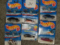 Seven Hot Wheels die cast cars in original 1998 First Editions packaging displayed on carpet. Models include Camaro Z28, Little Rattler, Ferrari F355 Barossa, and others.