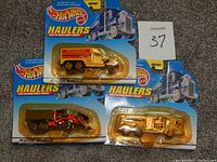 Three Hot Wheels Haulers diecast toy trucks in original packaging placed on carpet, showing front view with packaging intact, labeled with maxsold 37.