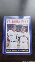 Photo of 1964 Topps baseball card featuring Willie McCovey and Leon Wagner titled 'Friendly Foes' in uniform, inside protective plastic sleeve.