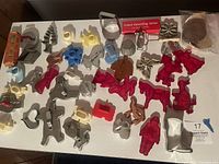 Wide shot showing the assorted cookie cutters spread out on a white surface, various materials and shapes visible including copper, aluminum, and colorful plastic cutters.