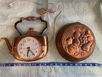 Copper finished electric teapot wall clock and round copper mold with embossed fruit design, both placed with a measuring tape for scale.