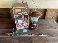 Photo of Coleman lantern, original box, and fishing rod lying on a wooden table showing complete items in the lot.