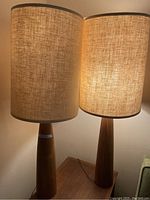 Pair of midcentury wood table lamps illuminated on a wood surface showing fabric shades and tapered wooden bases.