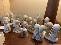 Collection of 11 Precious Moments porcelain figurines displayed on wooden surfaces, showing group arrangement with visible small dog detached from its base.