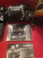 Photo of Canon black leather camera case with visible wear and two instruction manuals (Canon AE-1 and Speedlite 155A).