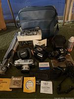 Photo showing two vintage cameras (Minolta silver and Nikon black), blue camera bag, tripod, two flashes, lens hoods, filters, cleaning paper, spray bottle and camera strap arranged on floor.