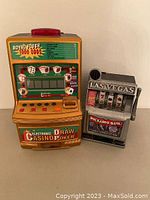 Photo of a gold-colored Electronic Casino Draw Poker slot machine toy and a silver Las Vegas Buckaroo Bank slot machine toy side by side.