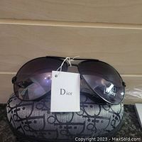 Front view of Dior sunglasses with tags beside the Dior-patterned case showing gradient lenses and metal frame.