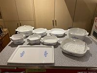 Full lot of vintage CorningWare baking and serving dishes arranged on a kitchen counter showing various sizes of casserole dishes with lids, rectangular serving platters with blue flower pattern.