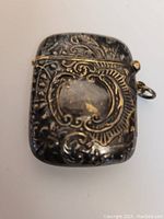 Close-up image of front side of silver match case showing detailed floral and scroll engraving, worn areas on surface.