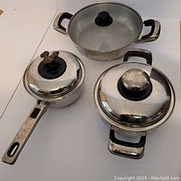 Six pieces of reflective stainless steel Carrera cookware arranged showing lids and handles. The set includes saucepans and pots with black handles and one glass lid.
