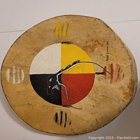 Top view of the drum face showing the hand painted four quadrant design with a black bird figure and colored sections in yellow, red, white, and black. Shows artist signature and date.