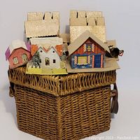 Top-down view showing all nine cardboard Christmas houses arranged inside the wicker basket. Glitter detail on rooftops visible.