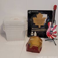 Photo showing all items including stack of clear plastic storage bins, Toronto Maple Leafs wooden paint by numbers kit in packaging, Iron Man mask, and guitar bank.