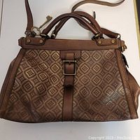 Front view of embossed leather satchel showing embossed pattern, buckle front detail, and leather straps.