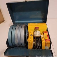 Blue metal tin case with the lid open showing five metal 5-inch movie tins with 8mm film reels, some film boxes and a bottle of film cement.