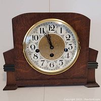 View of the front of the wooden mantle clock with clock face showing about 11:55. Clock dial with black Arabic numerals and gold center pattern. Clock face covered by brass bezel and glass door.