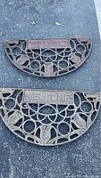 Two half-circle cast iron welcome mats placed on asphalt ground, showing overall design and size.