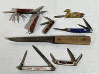 Collection of seven pocket-sized cutting tools including folding pliers multi-tool, novelty duck knife, various folding knives, and a large kitchen knife with wood handle