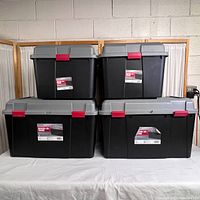 Four black storage containers with gray hinged lids and red locking clips, set on a white surface against a backdrop with wood frames and curtains.