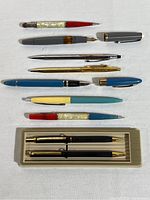 Full collection of 9 different pens and pencils arranged on white fabric, showing variety in style and color