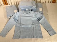 Five packages of light blue child-sized long sleeve T-shirts; each pack contains 5 shirts; one shirt unfolded and displayed to show shape and color.
