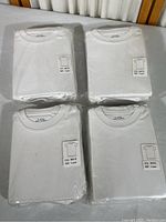 Four clear plastic packs each containing 5 white t-shirts lying flat on a surface. Each pack has a label reading COL: WHITE SIZE: 4 years.