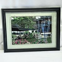 Full view of the framed reflective embroidery artwork displaying an Asian garden scene, showing the black wooden frame and reflection on glass.