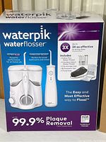 Front view of Waterpik Ultra Plus and Cordless Select water flossers on product box showing product image and features including plaque removal claims and included accessories.