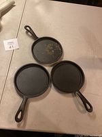 Lot of three cast iron pans including two flat griddle-style and one skillet with higher edges, shown on a table.