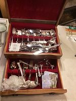 Top drawer of wooden flatware box containing forks, knives, spoons, and two serving ladles wrapped in plastic.
