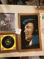 Framed portrait painting of Elvis Presley side profile with wooden frame next to framed vinyl and photo.