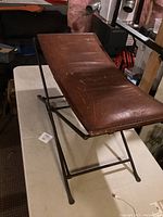 Full view of vintage brown leather foyer bench seat with angular cast iron folding legs on white table.