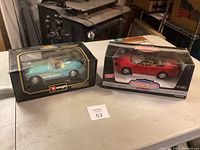 Two boxed model cars on table: a light blue Bburago Chevrolet Corvette and a red American Muscle convertible.