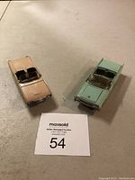 Two vintage model cars on a beige surface next to a MaxSold auction lot number 54 card.