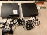Two Sony PS3 consoles side by side with controllers and cables on a flat surface, showing lot contents.