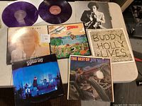 Seven vinyl records displayed on a table: Two purple vinyls from 'A War on Everything', and five black vinyls including Buddy Holly Lives, Joe Walsh, James Gang Live, Harry Nilsson and one partially obscured album.