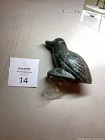 Top-down view of green soapstone bird carving on flat surface with auction tag.