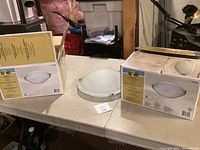 Three new Hampton Bay flush mount ceiling lights placed on a table, two in boxes and one unpackaged dome glass fixture.