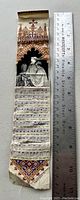 Full view of antique woven silk bookmark beside ruler for size reference showing intricate designs and musical notation.