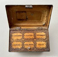 Antique metal spice box opened showing six labeled compartments inside the lid: cinnamon, cloves, nutmeg, ginger, allspice, mace