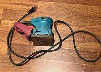 Makita finishing palm sander laid on wooden surface showing power cord with plug and attached dust collection bag.
