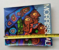 Front cover of the book showing colorful Indigenous-inspired artwork and the title 'Morrisseau'. A tape measure indicates book width about 12 inches.