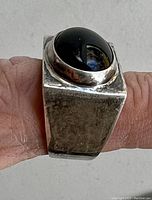 Front and side view showing large oval black stone bezel set on wide sterling silver band with textured finish.