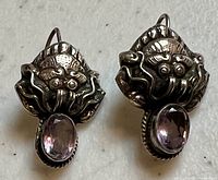 Pair of unique sterling silver earrings with a face or mask design and oval purple amethyst stones in bezel settings. Earring hooks visible for pierced ears.