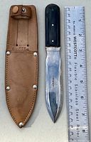 Cutting tool next to a ruler showing approximately 7 inches blade length, leather sheath with snap closure and riveted edges.