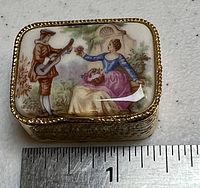 Photo of the closed porcelain pill box showing the Limoges style painted top of a man with a guitar and a woman with flowers, with a measuring ruler for size reference.